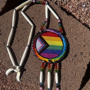 May include: A beaded necklace with a round, rainbow-colored pendant. The pendant is surrounded by a black beaded border and has a white, brown, and blue triangle in the center. The necklace is made of white bone beads and has a leather cord.