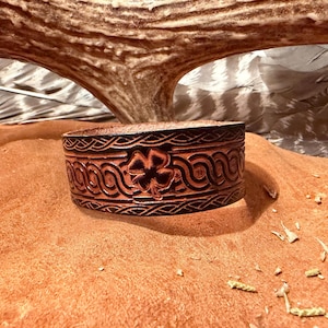 May include: A brown leather cuff bracelet with a carved Celtic knot and shamrock design. The bracelet is approximately 1.5 inches wide and features intricate detailing. The cuff is displayed on a tan surface.