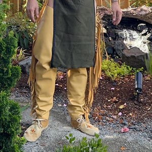 May include: A person wearing a traditional Native American outfit. The outfit includes tan leather pants with fringe, a dark green apron, and moccasins. The moccasins have decorative beadwork. The background features a garden setting with greenery and a wooden fence.