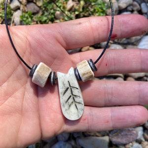 May include: A white bone necklace with a carved tree pendant and black beads. The necklace is on a black cord.