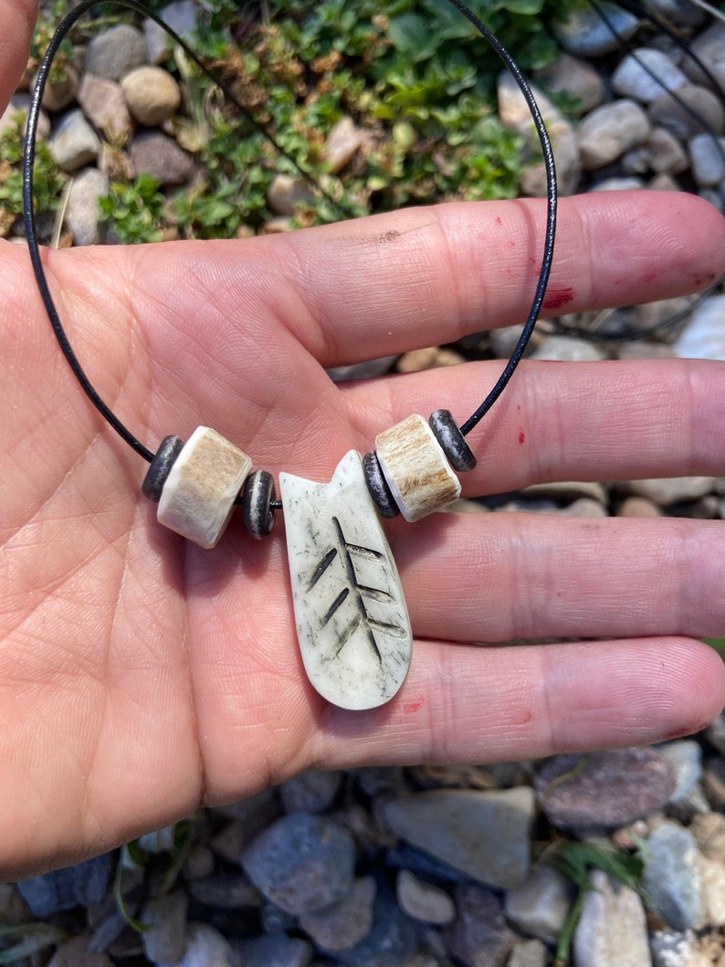 May include: A white bone necklace with a carved tree pendant and black beads. The necklace is on a black cord.