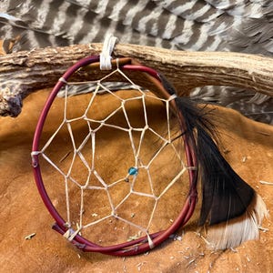 May include: A handcrafted dreamcatcher with a burgundy hoop and a woven web design. A black and brown feather is attached to the hoop. A small blue bead is in the center of the web. The dreamcatcher is displayed on a brown leather surface.