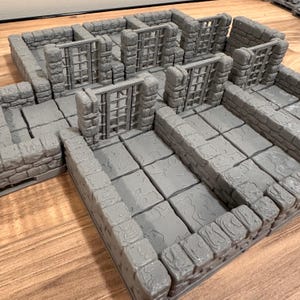 May include: A gray, modular dungeon terrain set with stone walls, floors, and barred gates. The set is designed for tabletop gaming, with multiple rooms and corridors. The pieces are textured to resemble aged stone.