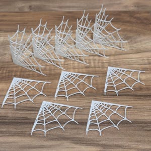 May include: A collection of white, corner-shaped spider web decorations. The webs are arranged on a wooden surface, with some in a corner shape and others flat. Ideal for Halloween or themed crafts.