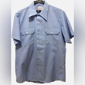 May include: Light blue short-sleeved shirt with two chest pockets and a button-down front. The shirt is hanging on a black hanger. The collar is a classic style.