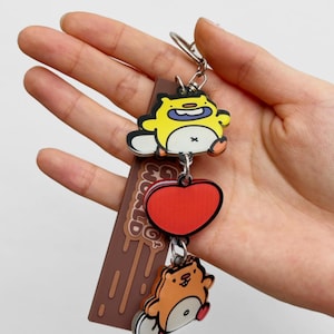 May include: A brown and white keychain with a chocolate bar design and the text "Go World Go". The keychain has three charms: a yellow cartoon character, a red heart, and a brown cartoon character.
