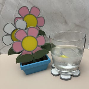 May include: A decorative flower arrangement in a blue pot with pink, white, and yellow flowers. A clear glass of water with ice sits on a flower-shaped coaster. The coaster is white with gray trim.