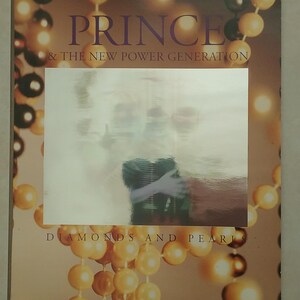 May include: A vintage poster for the album "Diamonds and Pearls" by Prince & The New Power Generation. The poster features a blurry image of Prince in the center, surrounded by a string of pearls.