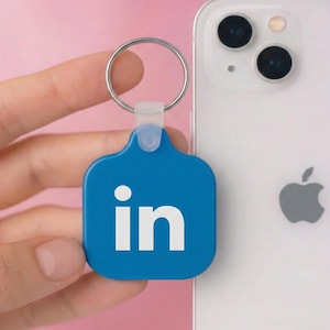May include: A square blue keychain featuring the white LinkedIn logo with the letters in. The keychain has a silver metal ring attached by a translucent plastic connector, positioned next to a white smartphone with a dual camera system.