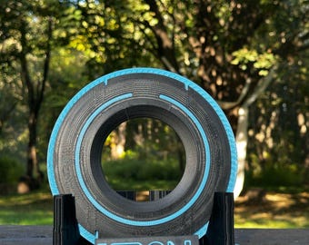 May include: A black and blue 3D-printed disc with a central opening, resting on a matching base. The word "TRON" is displayed on the base in blue. The disc has a blue illuminated ring around the outer edge.