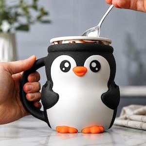 Puede incluir: Una taza de cerámica con forma de pingüino, con asa negra, parte superior negra y cuerpo blanco. El pingüino tiene ojos grandes y redondos, pico naranja y pies naranjas. La taza está llena de una bebida oscura y cubierta con una tapa blanca.