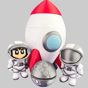 May include: A white and red rocket ship toy with a blue circular window, accompanied by two small astronaut figurines in white and silver suits and a textured silver moon sphere with visible craters positioned in the center foreground.