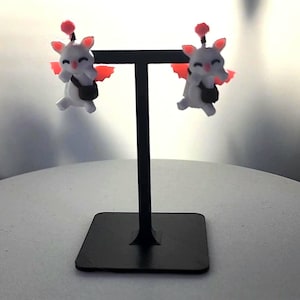 Op de afbeelding: Een paar wit en zwarte oorbellen met een stripfiguur met rode vleugels en een kleine zwarte tas. De oorbellen worden gepresenteerd op een zwarte metalen standaard met een vierkante basis.
