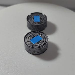 Op de afbeelding: Twee kleine, cilindervormige objecten met een spiraalvormig ontwerp en een felblauw vierkant in het midden. De objecten zijn donkergrijs en lijken van metaal te zijn. Ze liggen op een wit oppervlak.