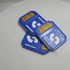 Puede incluir: Tres fichas rectangulares azules con las palabras "SUPER CREDIT" y un logotipo de flecha blanca. La parte superior de cada ficha tiene un diseño de código de barras de color dorado. Las fichas están apiladas unas sobre otras.