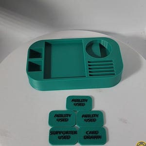 May include: A teal-colored tabletop organizer with various compartments, including a circular holder and several slots. Below are teal rectangular tokens with the text "ABILITY USED", "SUPPORTER USED", and "CARD DRAWN".