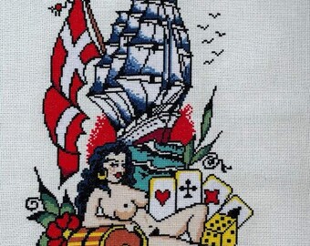 May include: A cross-stitch embroidery depicting a traditional nautical tattoo design. The design features a ship with a red, white, and blue flag, a woman with long black hair, playing cards, dice, and a red rose. The artist's signature "Tato - Jim" is visible in the lower right corner.
