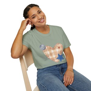 May include: A woman wearing a sage green t-shirt with a patchwork heart design. The heart is made of beige and white checked fabric with a blue star and a grey button.