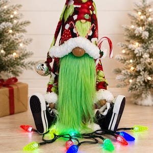 May include: A Christmas gnome figurine featuring a tall red hat with a holiday pattern, a long bright green beard, and a round wooden nose. The gnome wears black and white sneakers, holds a candy cane, and has a silver bell attached to its hat.