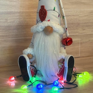May include: A plush gnome figurine wearing a white hat patterned with colorful Christmas light bulbs and a red glittery bulb accent. The gnome has a long white beard, wears red sneakers, and is surrounded by a string of glowing multi-colored lights.