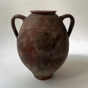 May include: A dark brown, weathered clay vessel with a rounded body, a narrow neck, and two symmetrical side handles. The surface features a mottled, textured finish with visible signs of age, wear, and uneven coloration across the exterior.