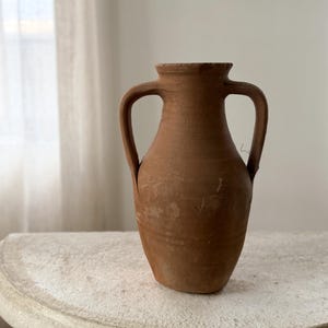 May include: A terracotta amphora vase with a matte, earthy brown finish. The vessel features a narrow neck, a wide rounded body, and two symmetrical handles attached to the sides. The surface shows natural texture and subtle variations in color.