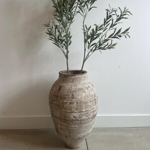 May include: A large, weathered, terracotta-colored ceramic jar with a textured, distressed white finish. The jar features horizontal ridges along its body and holds two tall, slender branches with green olive leaves and small dark olives.