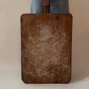 May include: A large, rectangular wooden cutting board with a weathered, distressed brown surface. The board features a central handle at the top, rounded corners, and a heavily worn, textured finish showing signs of age and extensive use.