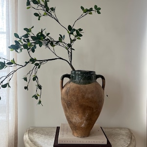 May include: A large, antique-style terracotta vase with a dark green rim and two handles. The vase holds a leafy branch. The vase sits on a stone-like table with two books titled "Arranging Things" and "Jake Arnold".