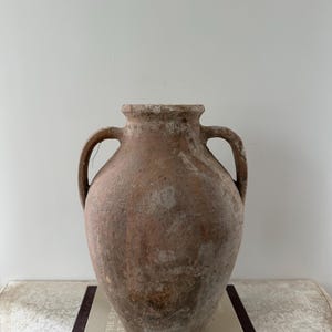 May include: A large, antique terracotta vase with two handles. The vase has a weathered, textured surface with shades of brown and beige. It sits atop a stack of books, suggesting a decorative or display item.