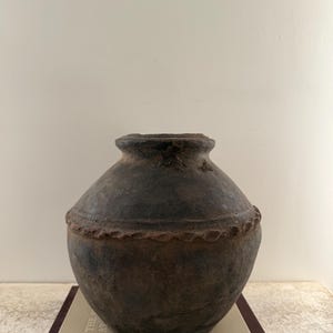 May include: A dark brown, antique-style ceramic vase with a rounded body and a textured surface. The vase has a decorative rim and sits on top of a book. The vase is likely used for home decor.