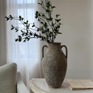 May include: A tall, weathered, brown ceramic amphora vase with two side handles, featuring a textured, aged finish. The vase contains a long, slender branch with small, dark green leaves extending upwards from the narrow opening at the top.