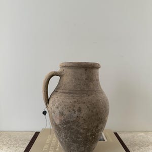 May include: A weathered, beige-colored ceramic jug with a handle. The jug has a rounded body and a slightly flared rim. It sits atop a book with the title "THE SPACE THAT KEEPS YOU". The jug has a tag attached to it.