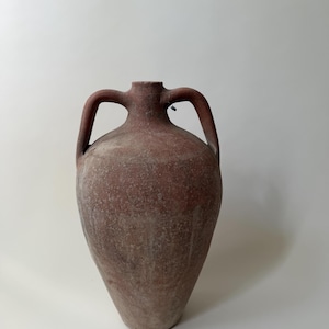 May include: A terracotta-colored earthenware amphora featuring a wide, rounded body that tapers toward the base, a narrow neck, and two symmetrical arched handles attached to the shoulders. The surface has a matte, weathered, and textured finish.