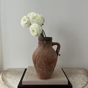 May include: A rustic, brown terracotta vase with a handle, holding a bouquet of white flowers. The vase sits atop two books with the title "ARRANGING THINGS" visible. The arrangement is on a light-colored, semi-circular table.