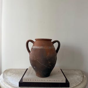 May include: A terracotta-colored amphora with two handles, displaying a weathered appearance with dark brown and black tones. The vase sits atop a stack of books with the title "Arranging Things" visible. The overall aesthetic suggests a vintage or antique decorative item.