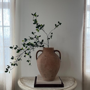 May include: A large, terracotta-colored vase with two handles, holding leafy green branches. The vase sits on a white, stone-like table, with two books stacked beneath it. The books have the title "ARRANGING THINGS" on the cover. Sheer curtains frame the background.