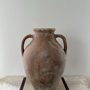 May include: A large, antique terracotta vase with two handles. The vase has a weathered, textured surface with shades of brown and beige. It sits atop a stack of books, suggesting a decorative or display item.