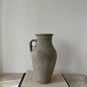 May include: A weathered, gray-green ceramic vase with a single handle. The vase has a wide body and a narrow neck. It sits atop two books, one light-colored with the text "ARRANGING THINGS" and a darker book with the text "JAKE ARNOLD".
