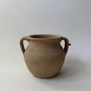 May include: A light brown, matte-finish earthenware pot with a wide, circular opening and two symmetrical, curved handles on opposite sides. The vessel features a rounded body and a smooth, unglazed surface with subtle natural texture variations.