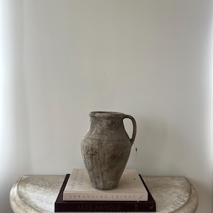 May include: A weathered, gray-brown ceramic vase with a single handle sits atop two books on a stone-like table. The books are titled "ARRANGING THINGS" and "JAKE ARNOLD". The vase has a rustic, aged appearance.