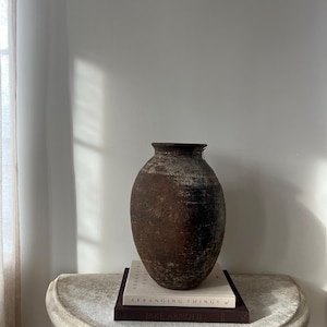 May include: A weathered, brown ceramic vase with a dark rim sits atop two books on a stone-like, semi-circular table. The vase has a textured surface and an oval shape. The books are titled "Arranging Things" and "Lake Arnold".