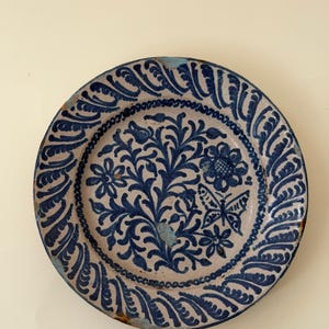 May include: A round ceramic plate with a cream-colored base and dark blue hand-painted designs. The center features a floral motif with a butterfly, while the rim is decorated with a repeating leaf pattern. The edges show signs of wear.