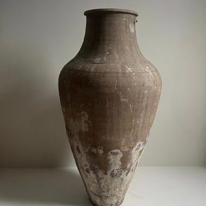 May include: A tall, tapered brown clay vessel with a wide, rounded body and a narrow neck. The surface features a weathered, matte texture with visible white mineral deposits and patches of discoloration concentrated toward the base of the object.