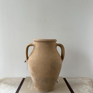 May include: A tan, terracotta-colored earthenware vessel featuring a wide, rounded body, a thick rim, and two symmetrical side handles. The surface has a matte, textured finish with subtle dark variations and a small tag attached to one handle.