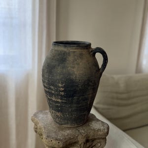 May include: A dark, weathered ceramic jug with a rounded body and a single side handle. The surface features a mottled, charcoal-black and earthy brown finish with visible horizontal ridges and a rough, aged texture throughout the exterior.
