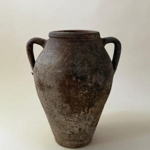 May include: A dark brown, weathered clay amphora with a wide, rounded body, a narrow neck, and two symmetrical side handles. The surface features a textured, mottled finish with visible signs of age, including light gray and earthy brown patches.