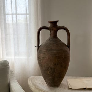May include: A tall, weathered brown clay amphora featuring a narrow neck, a flared rim, and two symmetrical arched handles extending from the neck to the upper body. The vessel has a textured, matte surface with visible signs of age and wear.