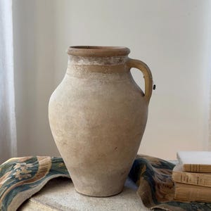 May include: A large, bulbous terracotta vessel with a wide mouth and a single side handle. The exterior features a weathered, matte beige finish with a darker, smooth tan band encircling the neck and the handle area, showing signs of age.
