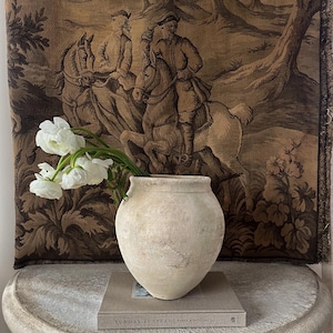 May include: A beige, textured ceramic vase holding white flowers, resting on a book titled "SUNDAY SUPPERS" on a round, stone-like table. A tapestry with a sepia-toned equestrian scene hangs in the background, creating a rustic, vintage aesthetic.